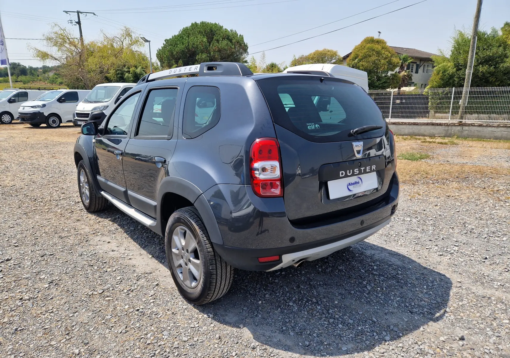 Vue 3/4 arrière droit d'un Dacia Duster gris foncé métal 2015 avec barres de toit et jantes alliage.