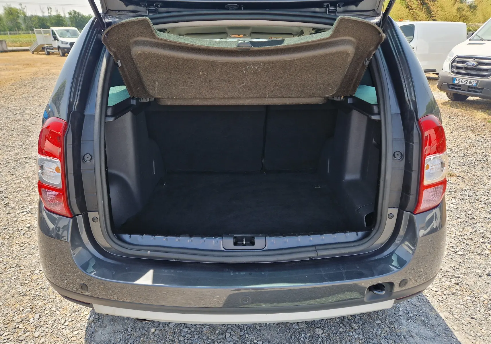 Vue arrière du coffre ouvert d'un Dacia Duster gris foncé métal, montrant l'espace de chargement vide et le hayon relevé.