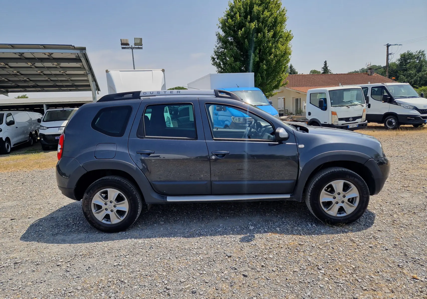 Profil côté droit d'un Dacia Duster gris foncé métal de 2015, avec barres de toit et jantes alliage visibles.