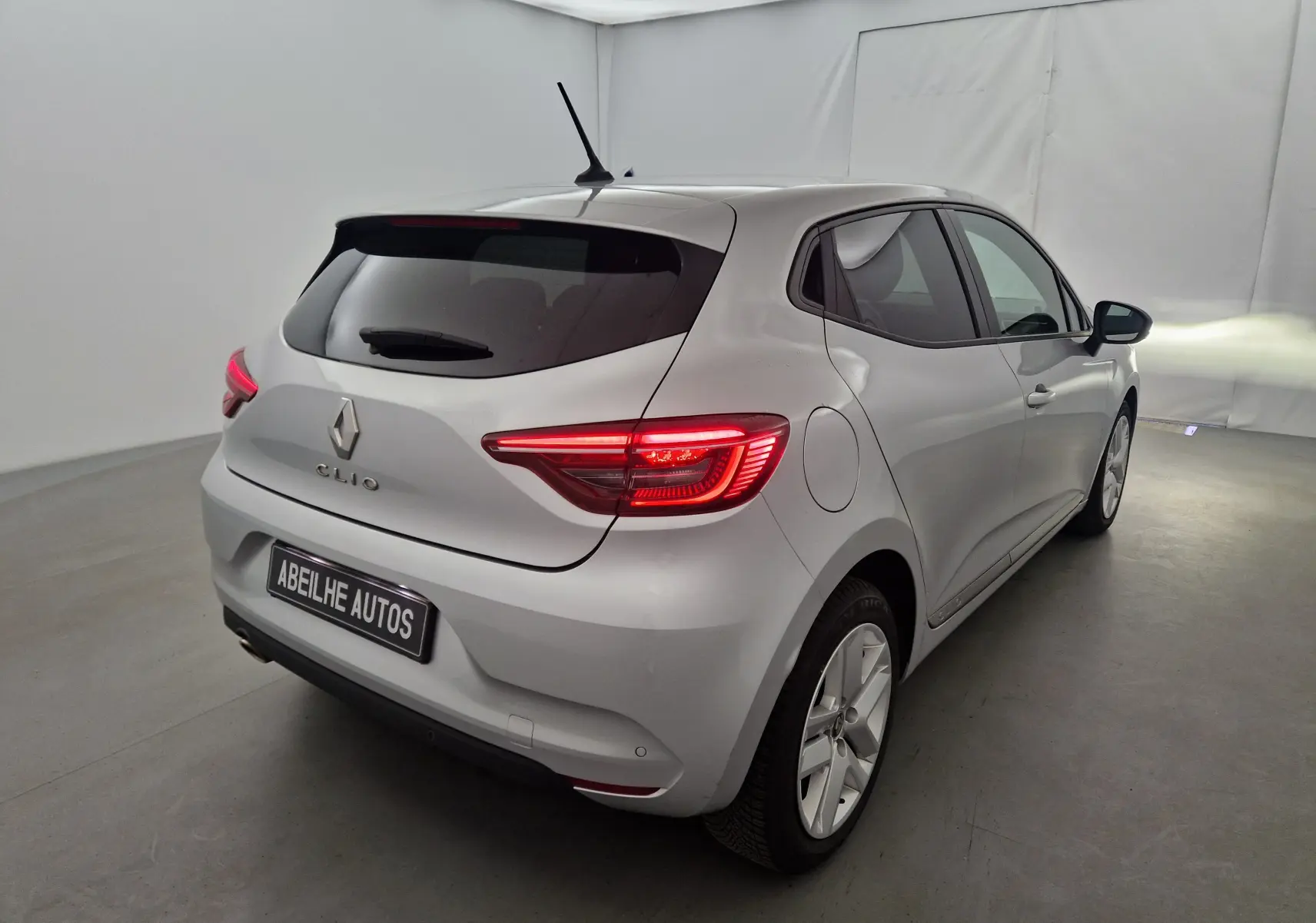 Renault Clio V gris platine vue 3/4 arrière droit dans un studio, feux arrière allumés et vitres teintées.