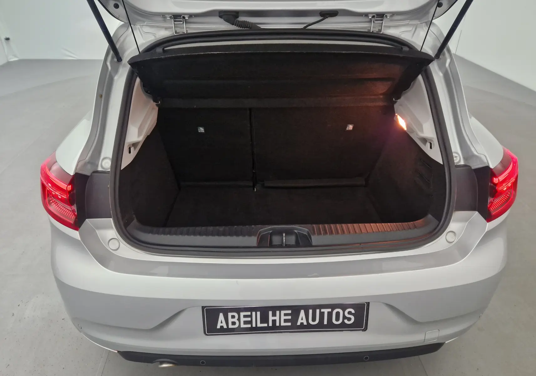 Coffre ouvert vu de l'arrière d'une Renault Clio V gris platine, avec feux arrière allumés et plaque Abeilhe Autos.