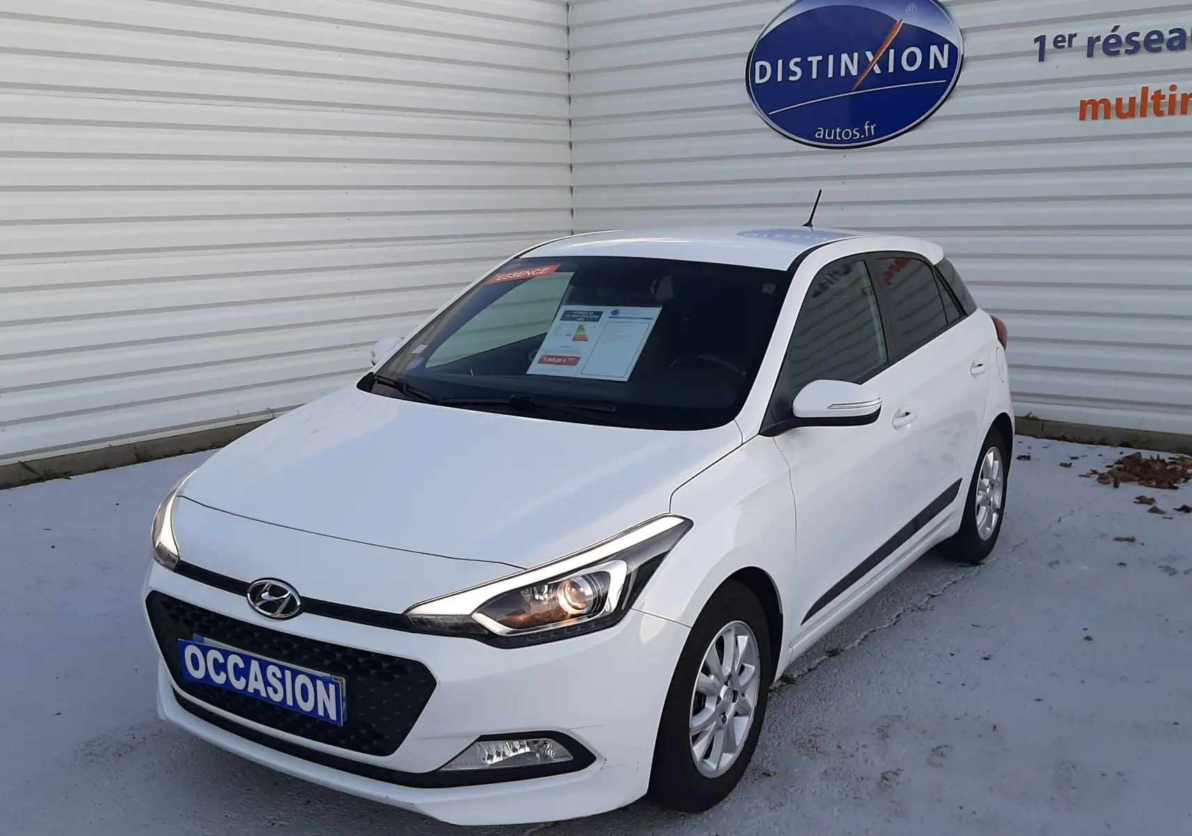 Vue 3/4 avant droite d'une Hyundai i20 blanche de 2016 avec phares allumés, stationnée devant un mur blanc.