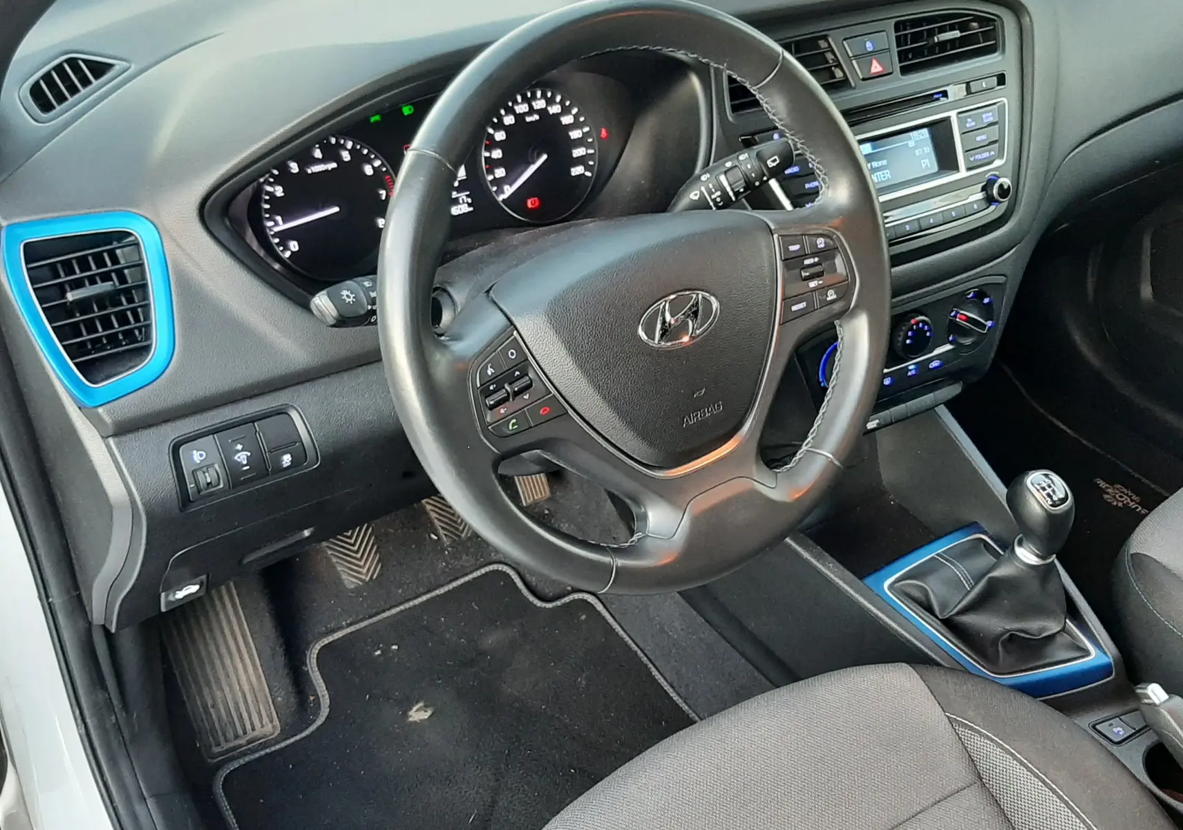 Vue intérieure côté conducteur de la Hyundai i20 blanche, volant multifonction et levier de vitesse manuel avec touches bleues.