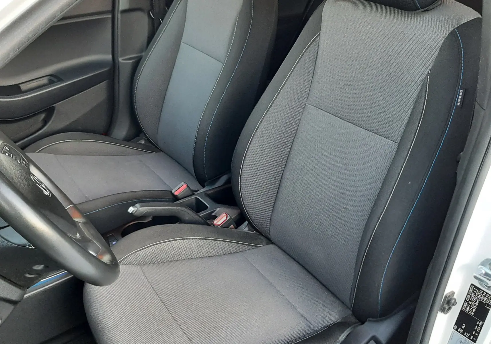 Vue intérieure côté conducteur de la Hyundai i20 blanche, sièges tissu gris avec logo i20 brodé en bleu sur les appuie-têtes.