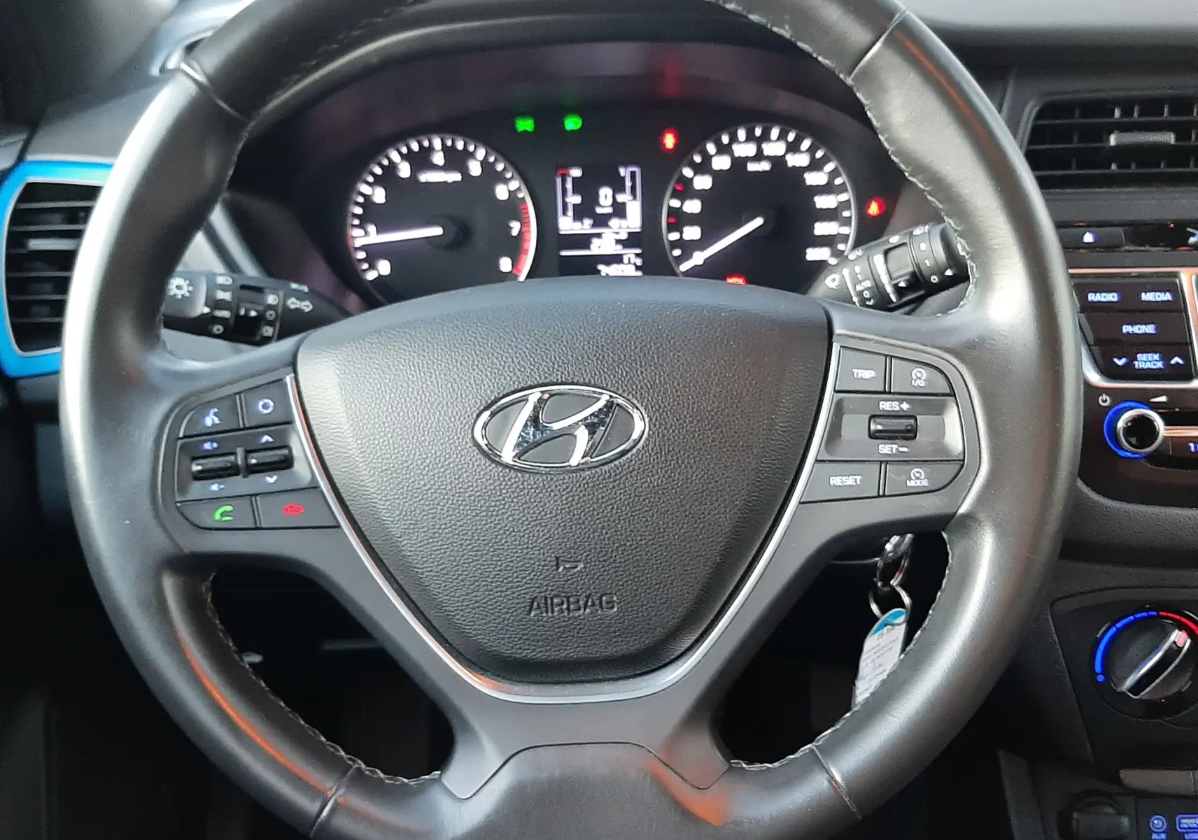 Vue rapprochée du volant noir de la Hyundai i20 2016 avec commandes intégrées et tableau de bord éclairé.