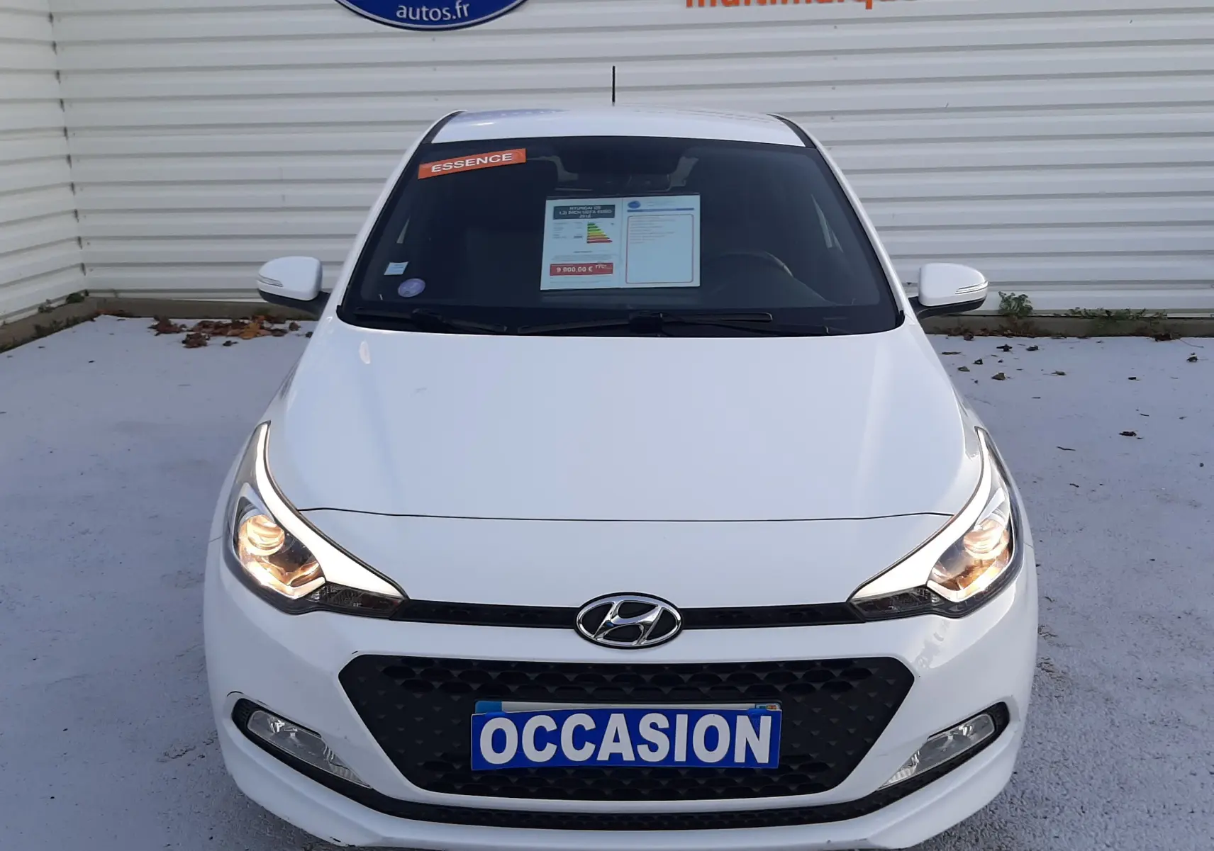 Vue avant d'une Hyundai i20 blanche de 2016 avec phares allumés et plaque "OCCASION" visible sur un parking.
