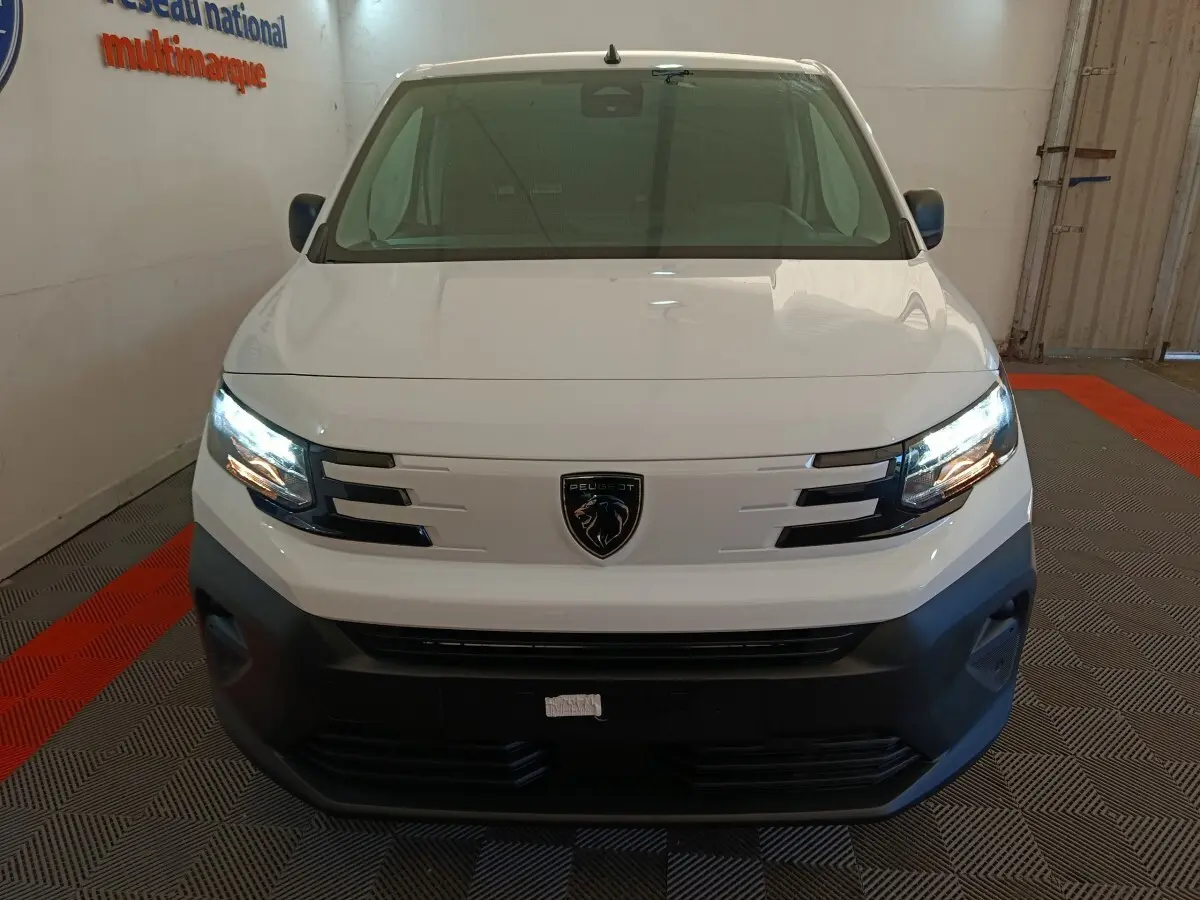 Vue frontale d'un Peugeot Partner blanc 2025 avec phares allumés et logo lion bien visible sur la calandre.
