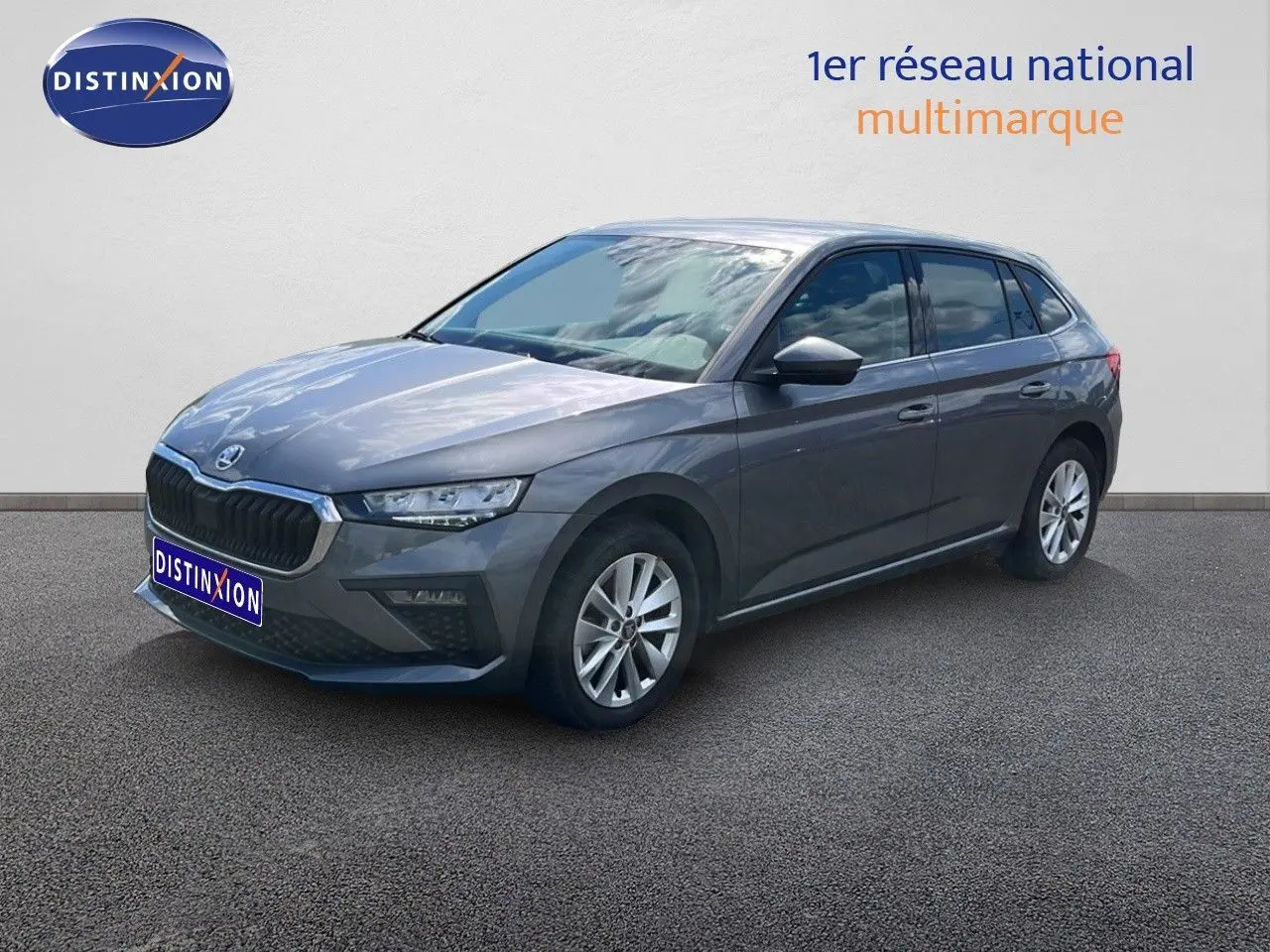 Vue 3/4 avant droite d'une Skoda Scala gris magnétique métallisé, stationnée devant un mur blanc avec logo Distinxion.
