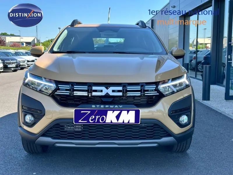 Vue frontale d'une Dacia Sandero Stepway beige Safari avec calandre noire et feux LED allumés sur parking.