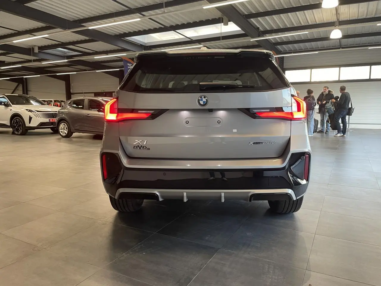 Vue arrière d'un BMW X1 xDrive 20d 2024 en gris Spacesilber Metallic avec feux arrière LED allumés en intérieur showroom.