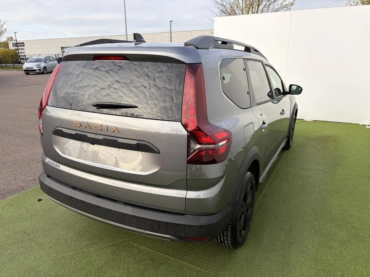 Vue 3/4 arrière droit du Dacia Jogger gris schiste métal avec barres de toit et logo Dacia brun cuivré visible.
