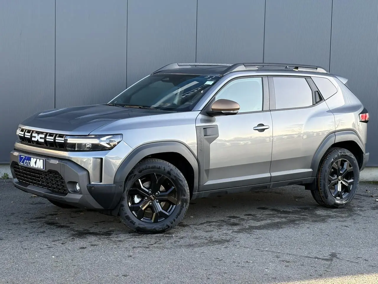 Vue 3/4 avant droit du Dacia Duster 2026 gris schiste avec jantes noires et barres de toit longitudinales.