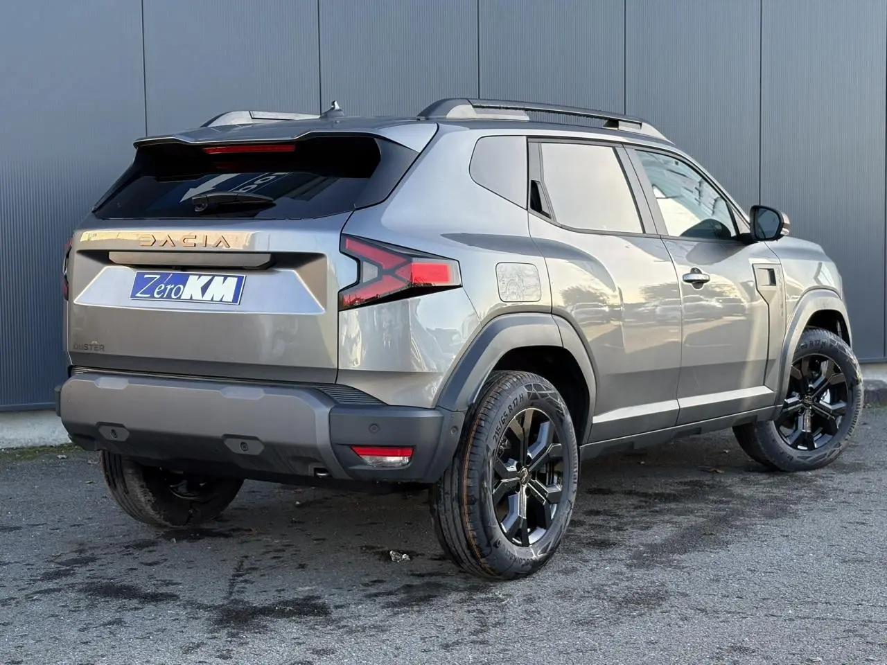 Vue 3/4 arrière droite du Dacia Duster 2026 gris schiste avec jantes alliage noires et barres de toit longitudinales.