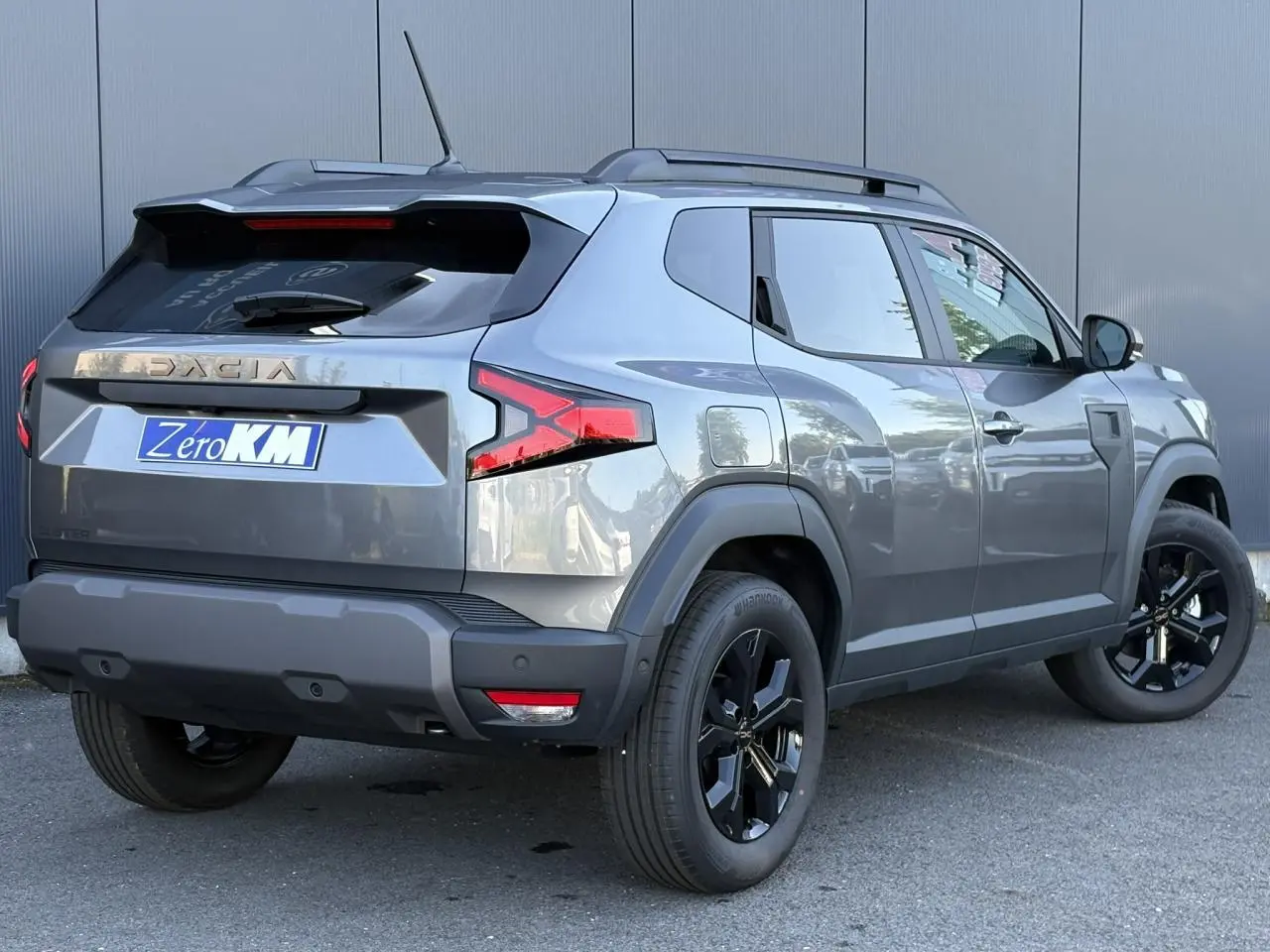 Vue 3/4 arrière droite du Dacia Duster 2026 gris schiste avec jantes alliage noires et barres de toit longitudinales.
