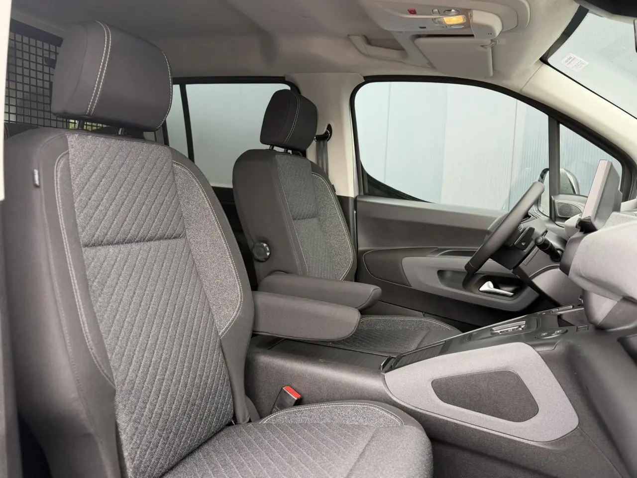 Vue intérieure côté conducteur du Peugeot Rifter gris acier 2026, sièges tissu gris avec accoudoirs et tablette tactile visible.