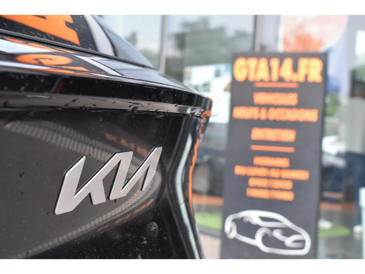 Gros plan sur le logo KIA à l'arrière d'un SUV noir, avec un panneau flou en arrière-plan.