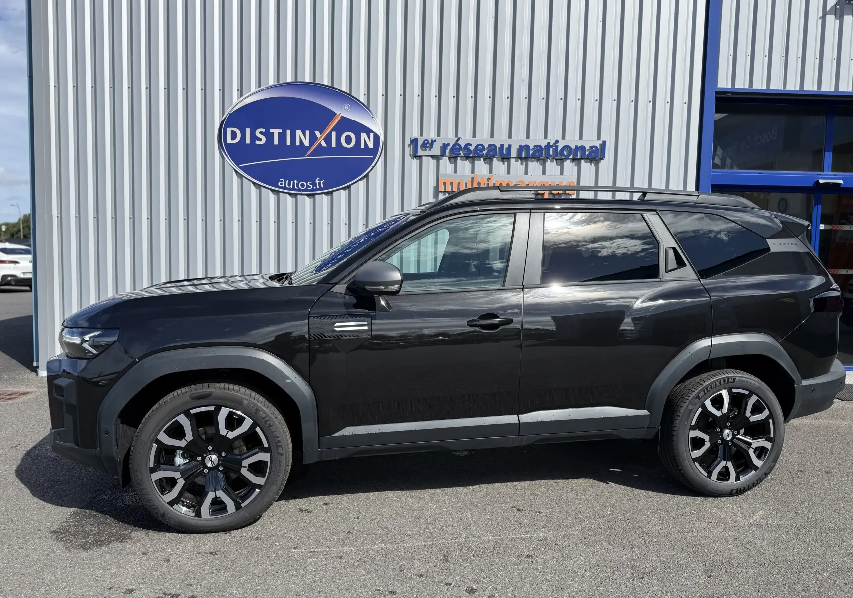 Profil droit du SUV Dacia Bigster Hybrid 155 Journey noir nacré avec jantes biton 19 pouces, garé devant un mur métallique.