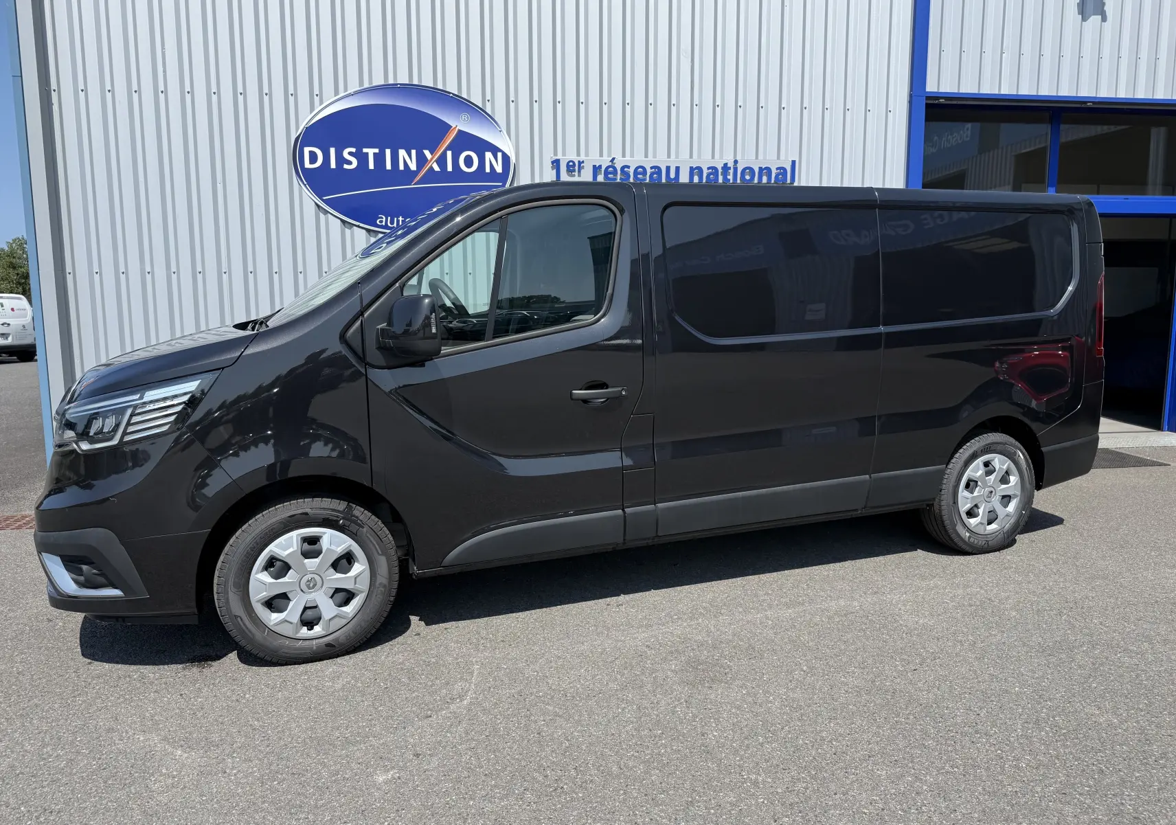 Renault Trafic Fourgon noir crépuscule métal vu de profil côté gauche, stationné devant un bâtiment commercial.