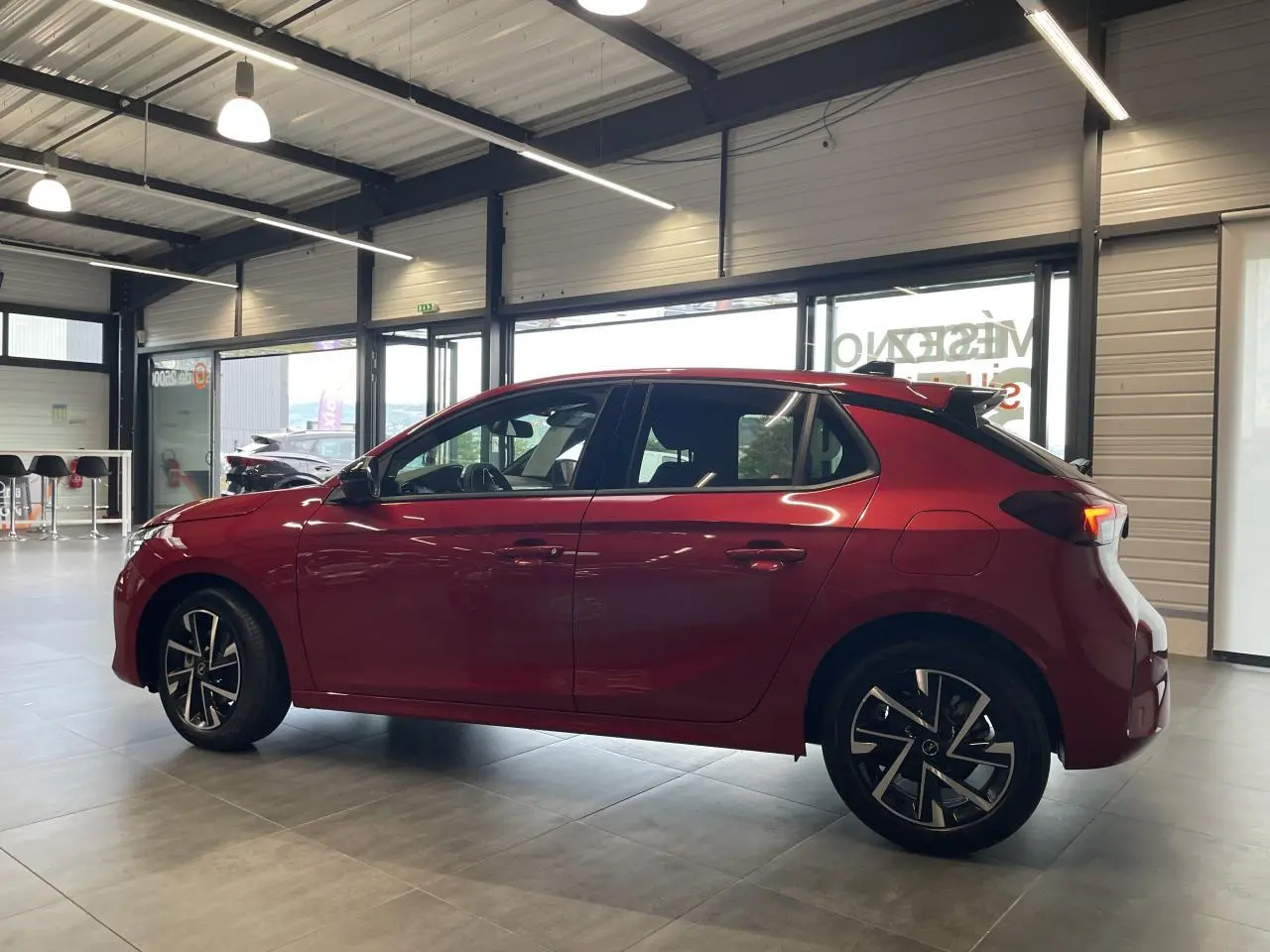 Opel Corsa rouge vue de profil côté gauche en intérieur, avec jantes alliage et toit couleur carrosserie.