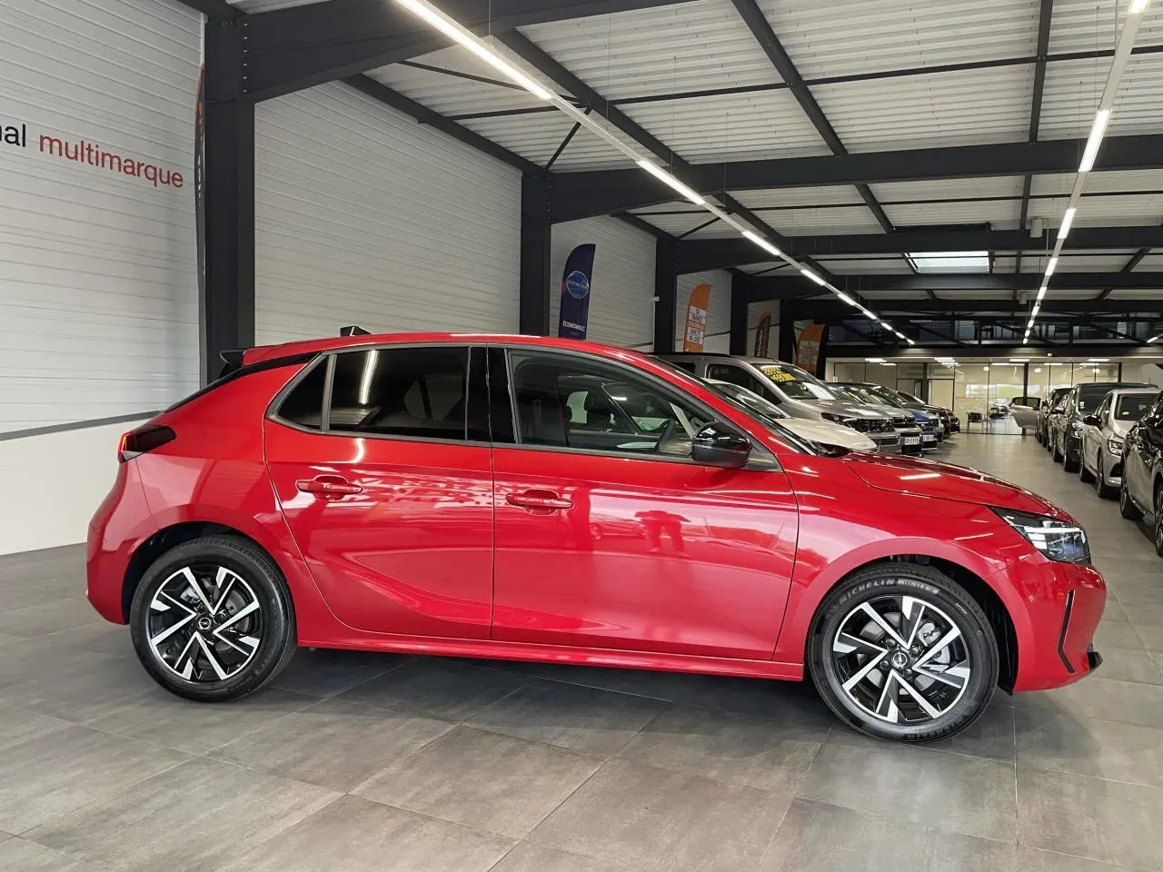 Opel Corsa rouge vue de profil côté gauche en intérieur, avec jantes alliage et vitres surteintées.