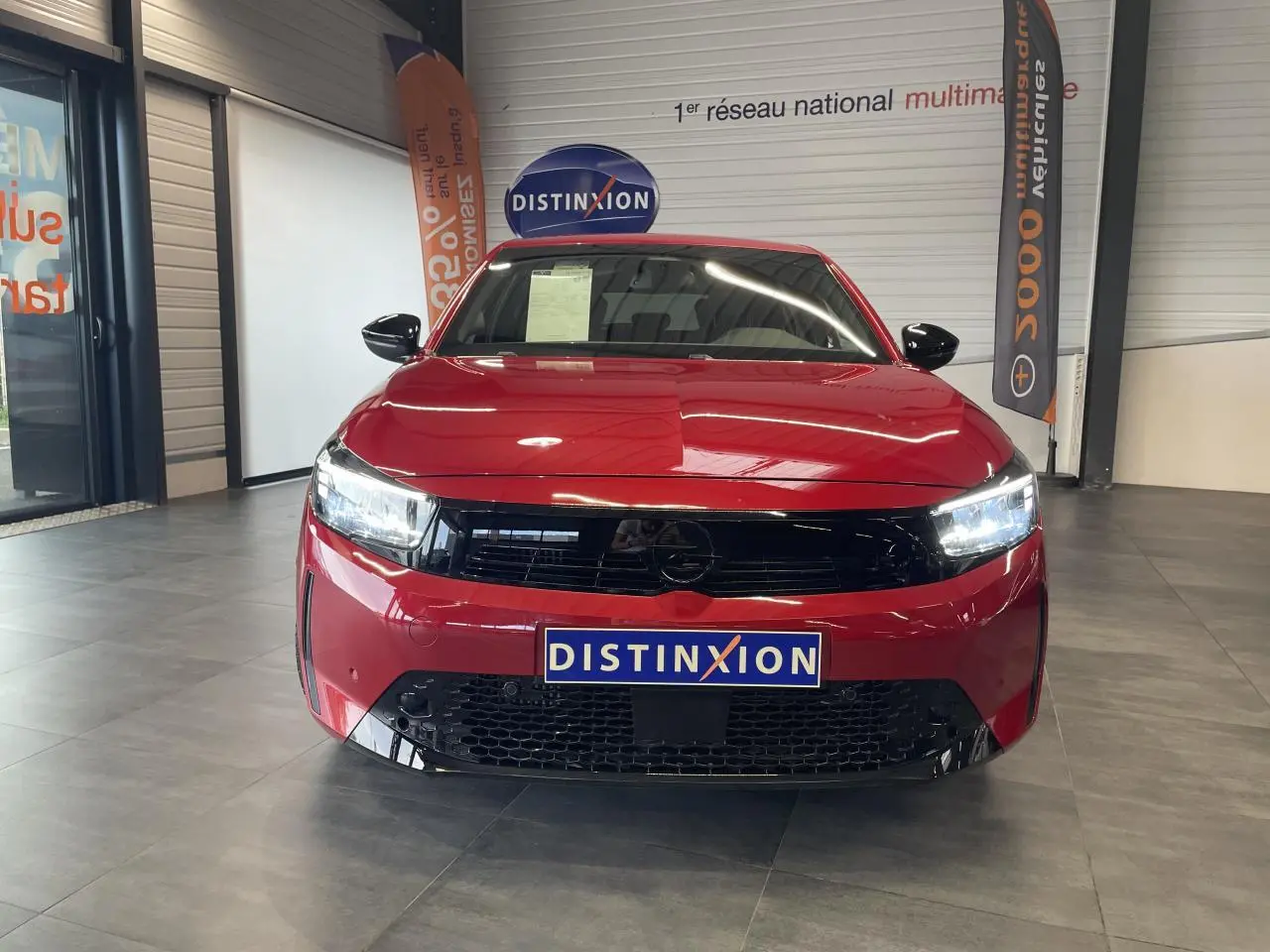 Vue avant d'une Opel Corsa rouge 2026 avec calandre noire brillante et phares LED allumés en intérieur showroom.