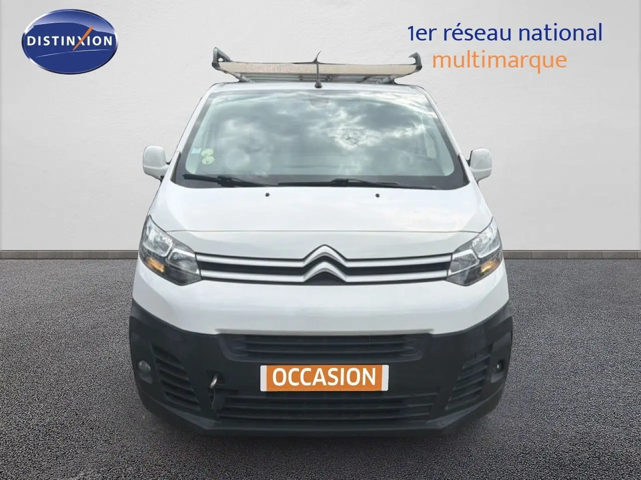 Vue avant d'un Citroën Jumpy XL blanc avec pare-chocs noir et galerie de toit, en arrière-plan un mur gris.