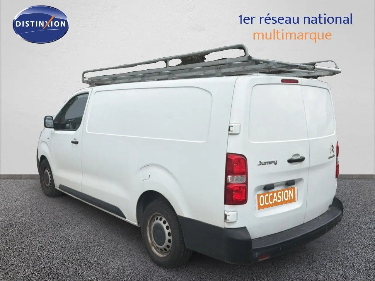Vue 3/4 arrière gauche d’un utilitaire Citroën Jumpy blanc avec galerie de toit et marquage « Occasion ».
