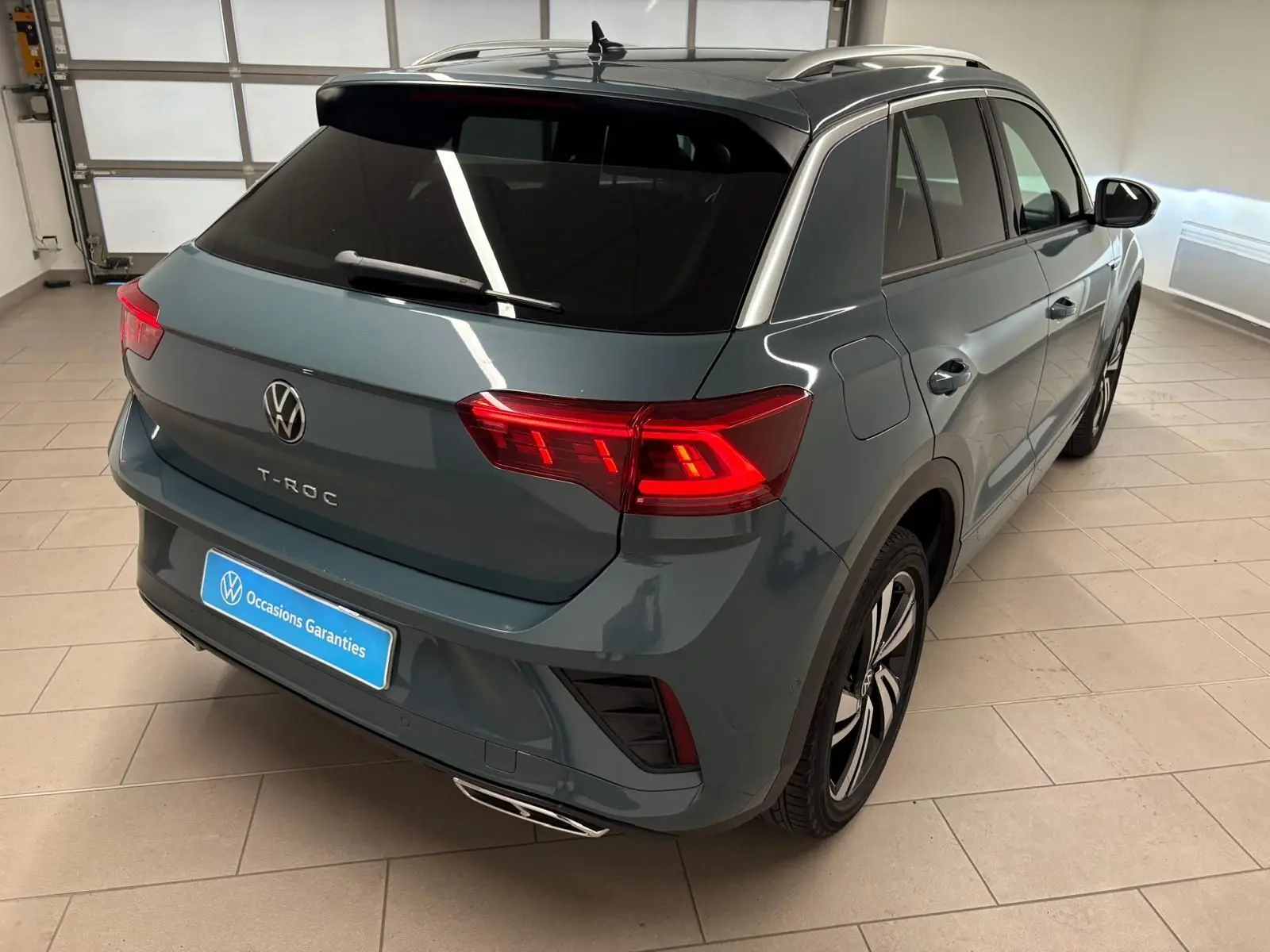 Vue 3/4 arrière droite du Volkswagen T-Roc 2024 bleu pétrole avec feux arrière LED et finition R-Line en intérieur showroom.