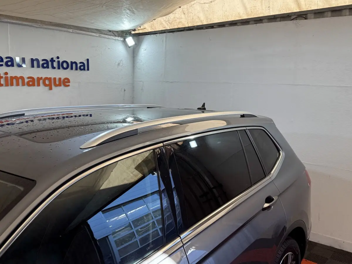 Vue 3/4 arrière côté droit d’un Volkswagen Tiguan Allspace gris foncé avec toit ouvrant et barres de toit chromées.