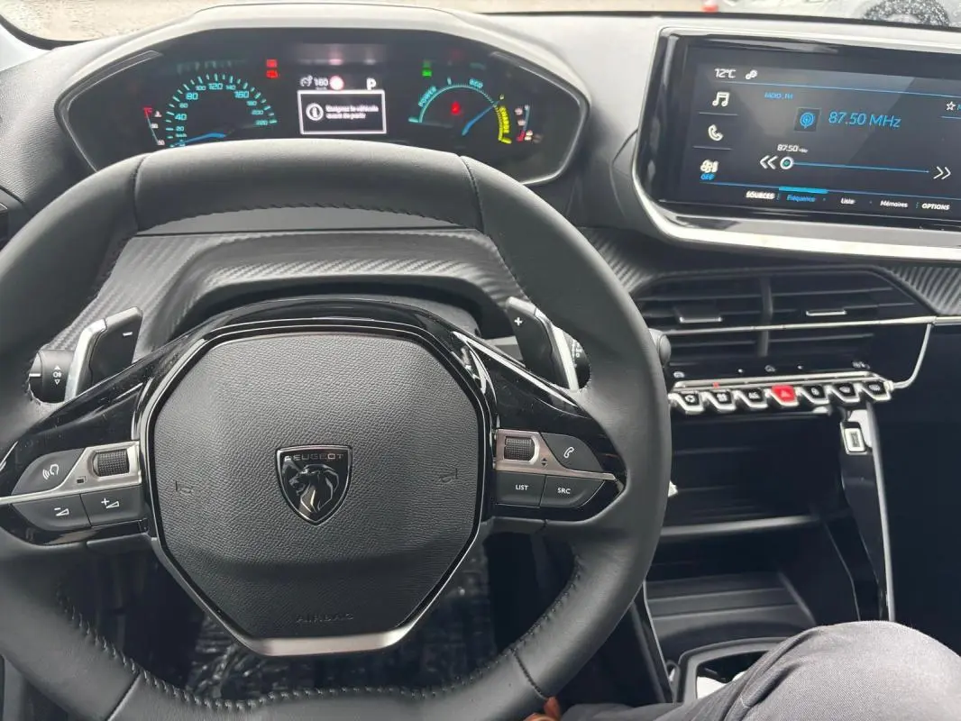 Vue intérieure centrée sur le volant Peugeot 208 gris, avec cockpit numérique et écran tactile radio visible.