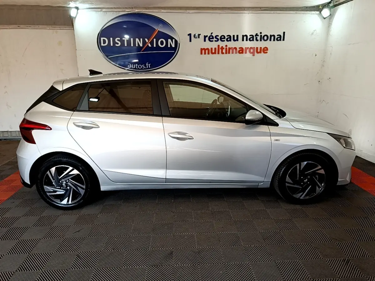 Vue de profil côté gauche d'une Hyundai i20 gris clair 2022 dans un showroom avec jantes noires et argentées.