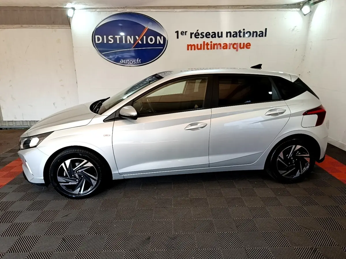 Vue de profil côté gauche d'une Hyundai i20 gris clair 2022 en intérieur avec jantes alliage noires et logo Distinxion en arrière-plan.