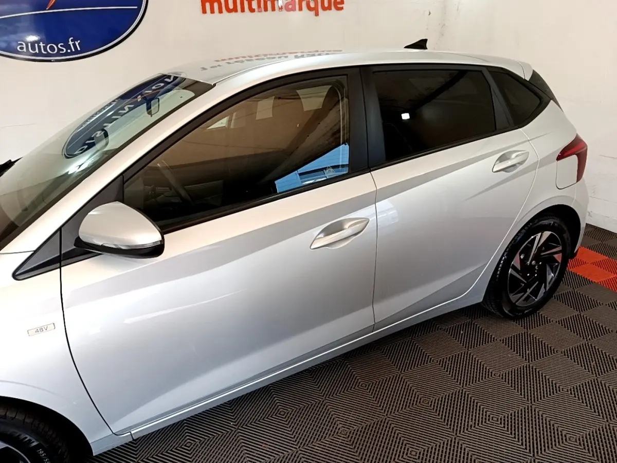 Vue côté gauche d'une Hyundai i20 gris clair 2022, mettant en valeur les jantes noires et les vitres teintées.