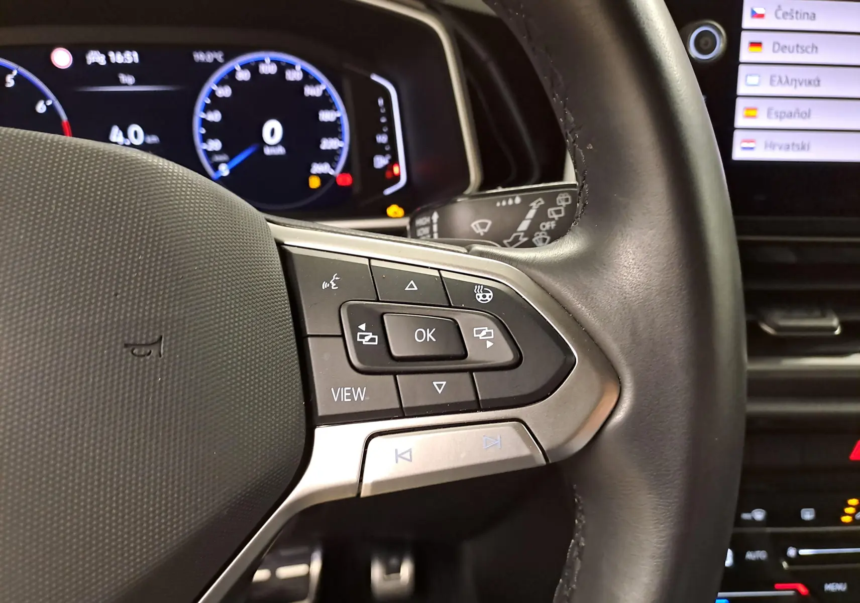 Gros plan sur les commandes du volant du Volkswagen T-Roc Gris Ascot, avec tableau de bord numérique en arrière-plan.