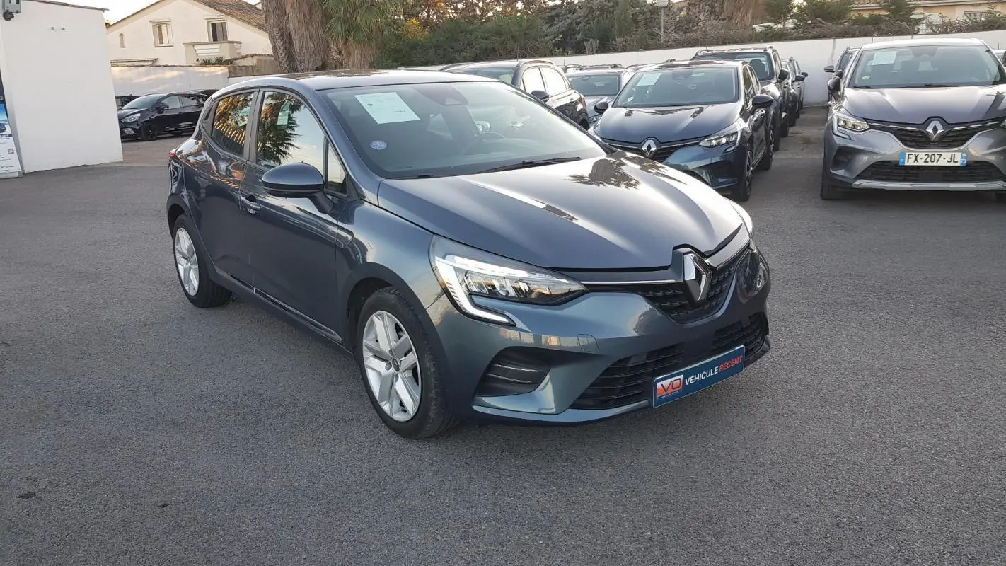 Renault Clio gris en vue 3/4 avant droit, avec phares allumés et jantes alliage visibles sur parking.