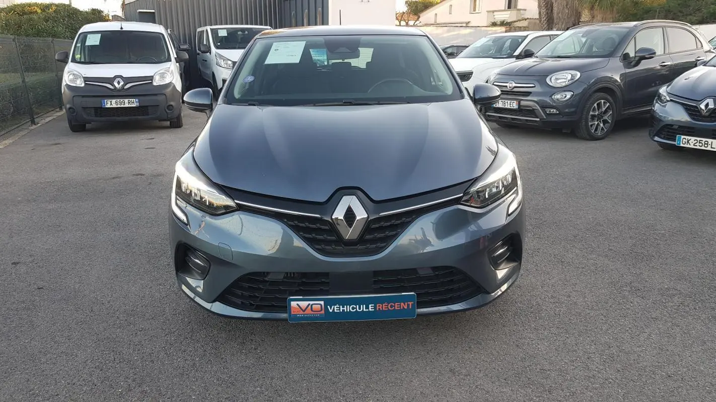 Vue frontale d'une Renault Clio gris métallisé avec feux allumés et plaque "Véhicule récent" sur un parking.