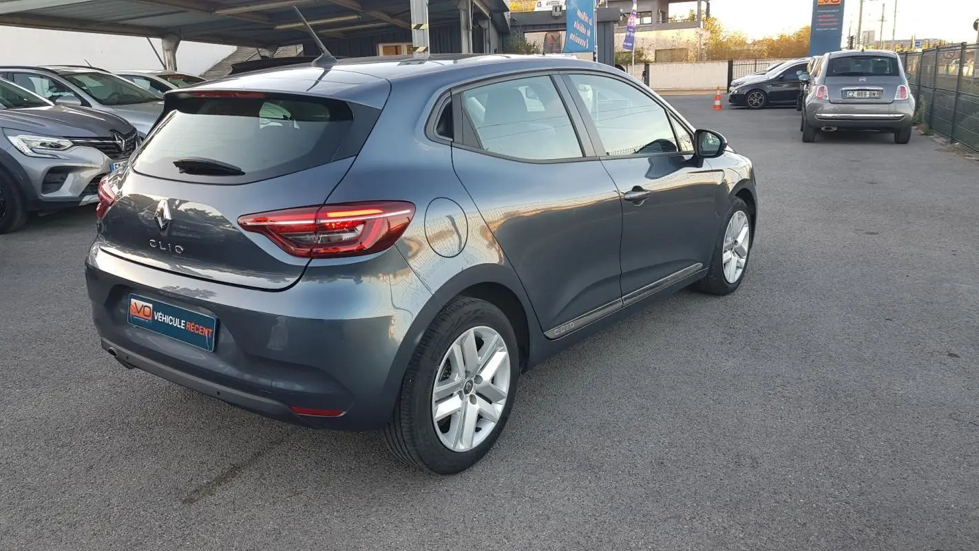 Renault Clio gris vue 3/4 arrière droit sur parking, avec feux arrière allumés et jantes alliage visibles.