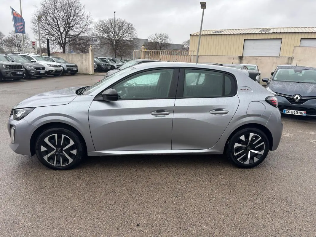 Vue de profil côté gauche d’une Peugeot 208 Hybrid 100 e-DCS6 Style gris avec jantes 16 pouces au parking sous la pluie.
