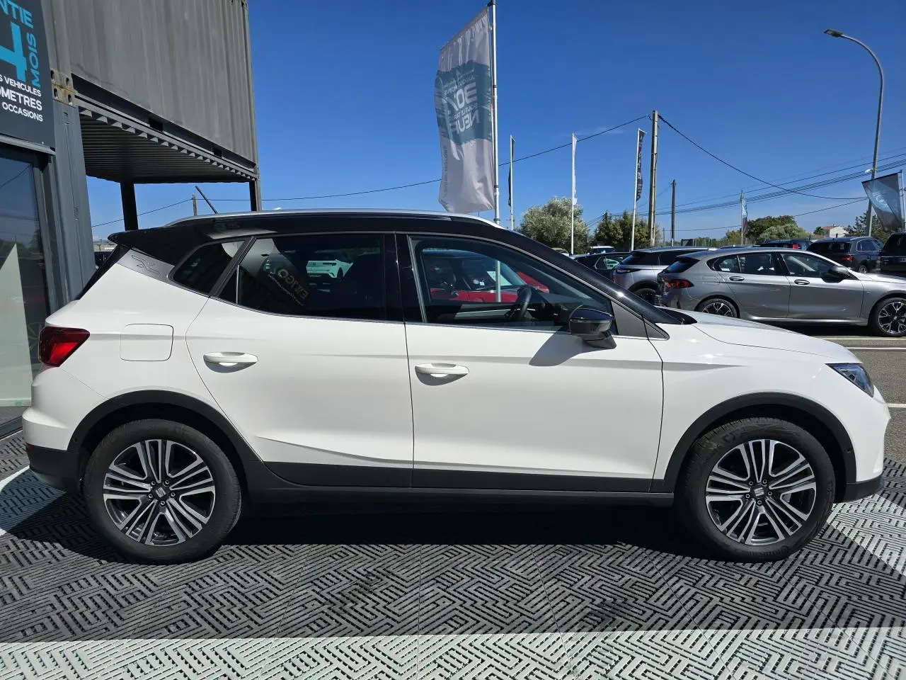 Vue de profil droit d'un SEAT Arona 2024 blanc avec toit noir et jantes alliage, garé en extérieur sous un ciel bleu.