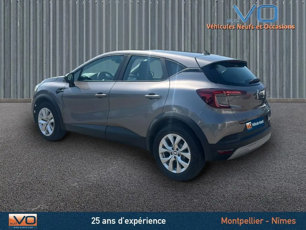 Renault Captur TCe 90 Business gris en 3/4 arrière droit, stationné devant un mur métallique.