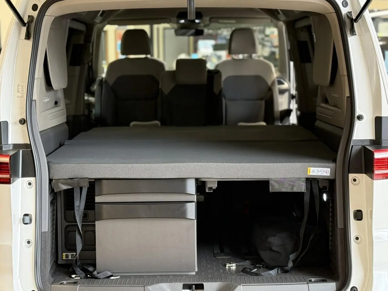 Vue arrière intérieure du Volkswagen California T7 blanc Candy avec couchette déployée et rangements sous le lit.