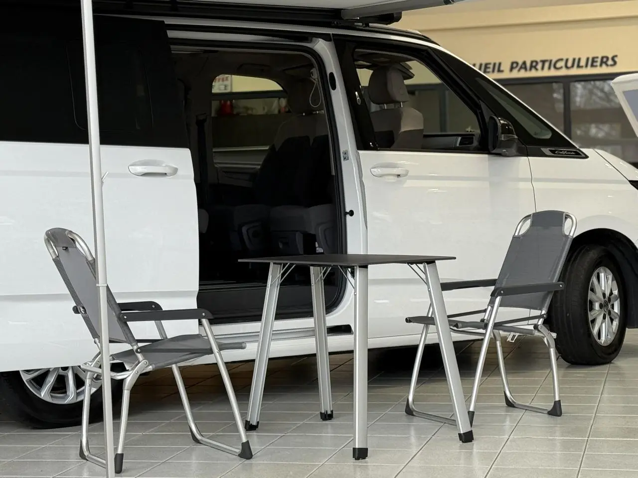 Vue latérale droite d’un Volkswagen California T7 blanc Candy avec porte coulissante ouverte et table de camping entre deux chaises.