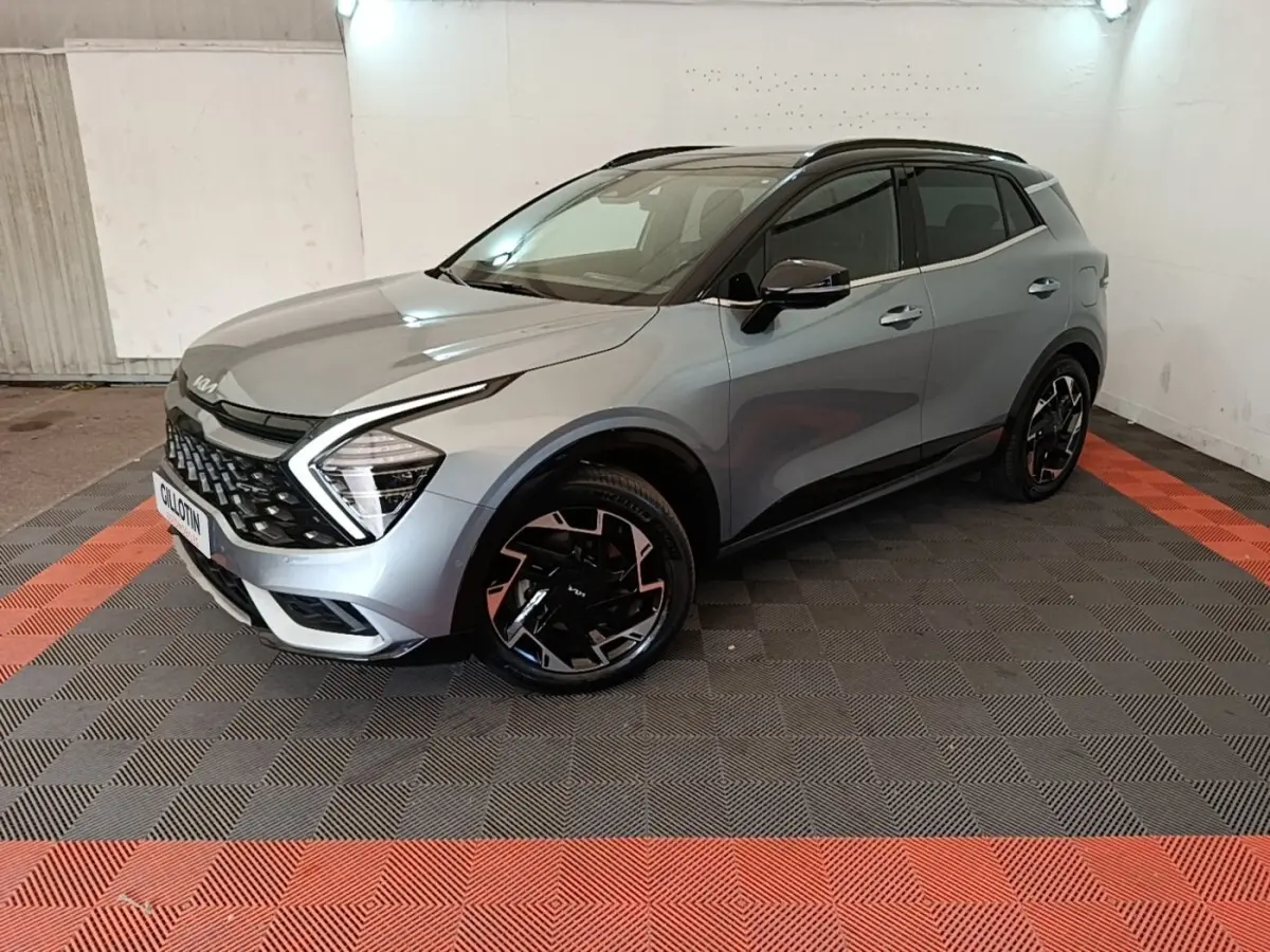 KIA Sportage gris clair en 3/4 avant droit, avec jantes noires et toit noir dans un garage éclairé.