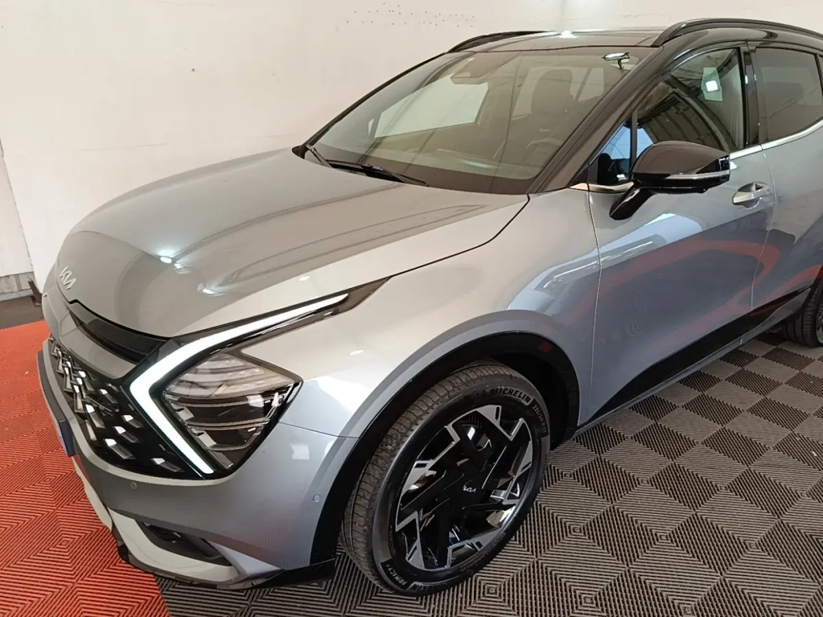 Vue 3/4 avant droit d’un KIA Sportage gris clair avec phares LED en forme de boomerang et jantes noires distinctives.
