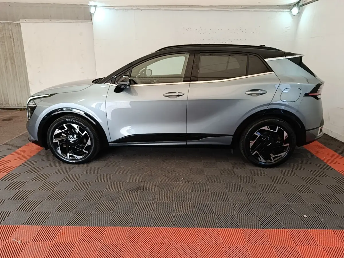 Vue de profil côté gauche d'un KIA Sportage gris clair avec toit noir et jantes noires stylisées, stationné en intérieur.