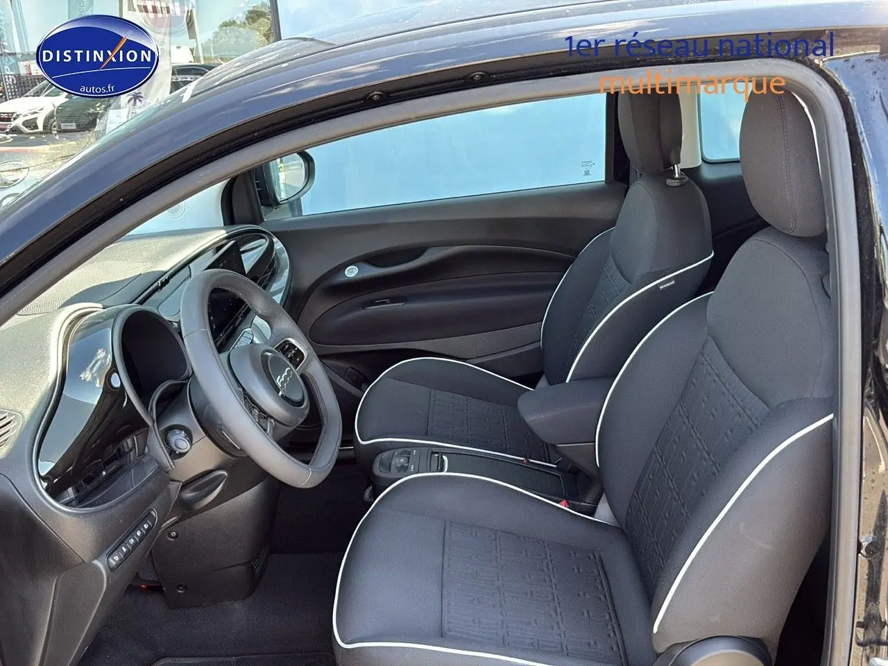 Intérieur de la Fiat 500 électrique 2024 vu côté gauche, sièges noirs avec surpiqûres blanches et volant multifonction.