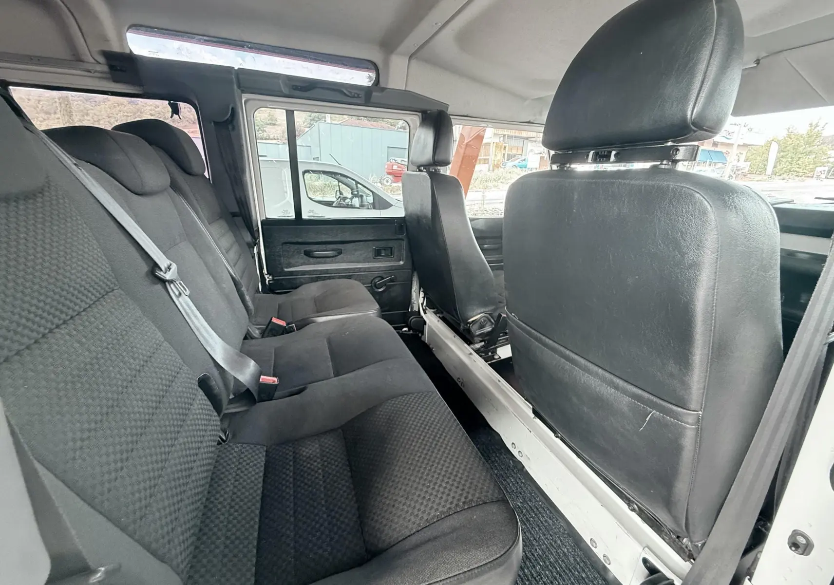 Vue intérieure côté gauche du Land Rover Defender 2007 blanc, montrant les sièges arrière en tissu gris noir et les sièges avant en cuir noir.