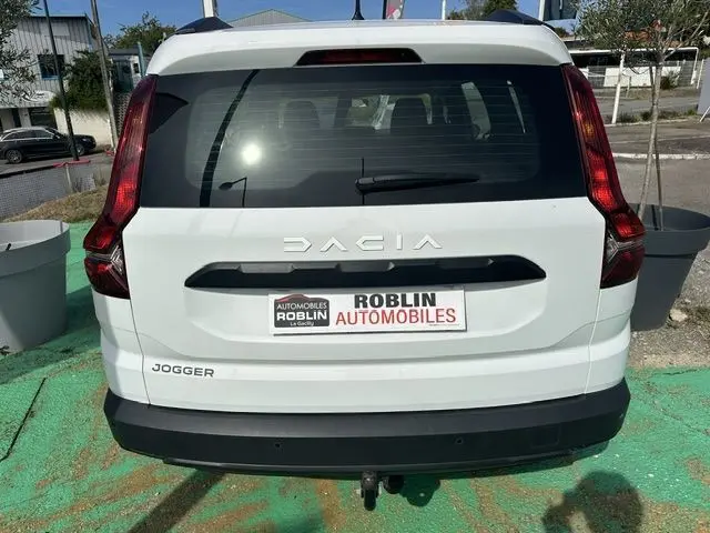 Vue arrière d'un Dacia Jogger blanc 2023 avec feux verticaux rouges et logo visible sur un parking extérieur.
