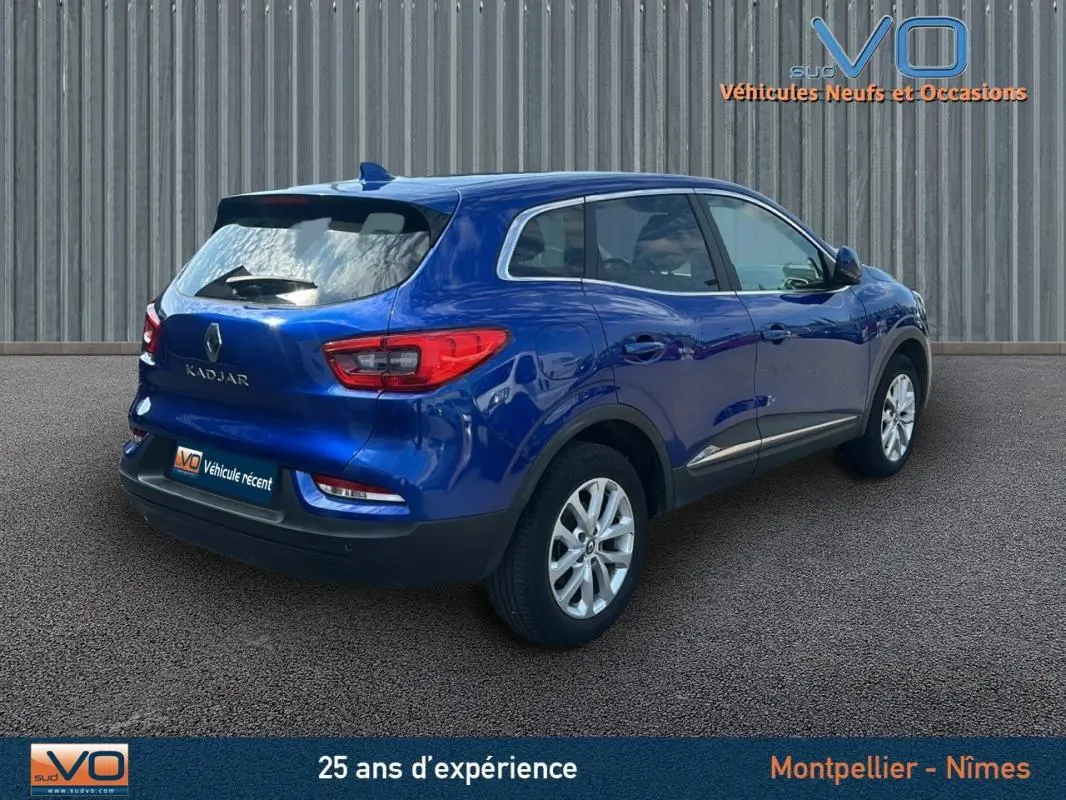 Vue 3/4 arrière droite d'un Renault Kadjar bleu foncé avec barres de toit et jantes alliage 17 pouces sur parking.