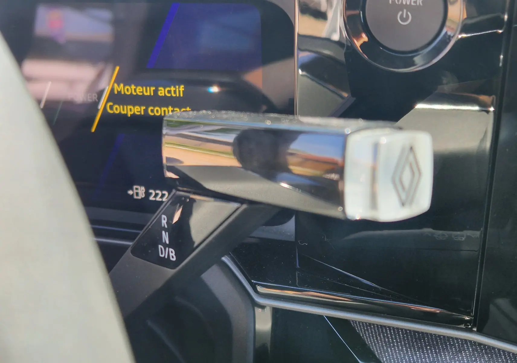 Gros plan sur le levier de boîte automatique chromé avec logo Renault, tableau de bord numérique en arrière-plan.